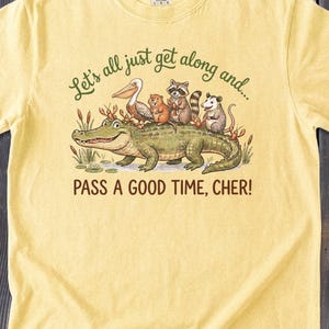 May include: A pale yellow short-sleeve t-shirt featuring a graphic of an alligator with a pelican, raccoon, opossum, and nutria on its back. The text reads: Let's all just get along and... PASS A GOOD TIME, CHER! The shirt has a crew neckline.