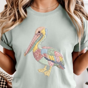 May include: A light sage green t-shirt featuring a central graphic of a pelican designed with a patchwork aesthetic. The bird is composed of various pastel-colored fabric patterns, including floral, gingham, and solid yellow, pink, and blue sections.