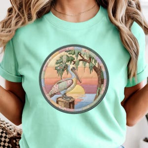 May include: A mint green t-shirt featuring a circular graphic print of a pelican standing on a wooden post. The design includes a sunset background with horizontal stripes, a tree with hanging moss, and a patchwork-style border in pastel colors.