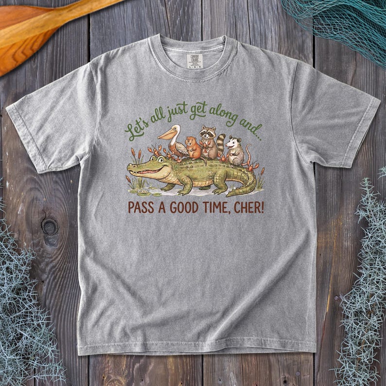 May include: A grey short-sleeve t-shirt featuring a graphic of a large green alligator with a pelican, raccoon, beaver, and opossum on its back. The text reads: Let's all just get along and... PASS A GOOD TIME, CHER! The shirt has a crew neckline.