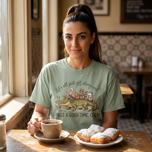 May include: A cream-colored short-sleeve t-shirt featuring a graphic of a green alligator carrying a pelican, a beaver, a raccoon, and an opossum. The text reads: Let's all just get along and... PASS A GOOD TIME, CHER! The design includes small crawfish.