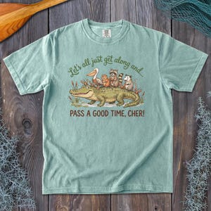 May include: A light green short-sleeve t-shirt featuring a graphic of an alligator, pelican, beaver, raccoon, and opossum. The text reads: Let's all just get along and... PASS A GOOD TIME, CHER! The shirt has a crew neckline and a relaxed fit.