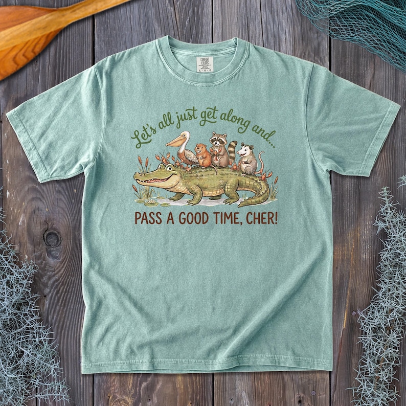 May include: A light green short-sleeve t-shirt featuring a graphic of an alligator, pelican, beaver, raccoon, and opossum. The text reads: Let's all just get along and... PASS A GOOD TIME, CHER! The shirt has a crew neckline and a relaxed fit.