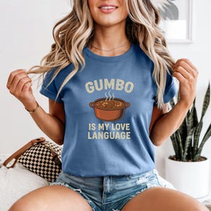 May include: A blue t-shirt with the text "Gumbo is my love language" and a graphic of a pot of gumbo. The text and graphic are in a vintage style. The shirt has short sleeves and a crew neck.