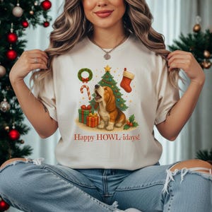 Puede incluir: Camiseta color crema con un basset hound aullando junto a un árbol de Navidad, regalos y un calcetín. La camiseta tiene el texto "Happy HOWL-idays!" impreso en rojo. El diseño incluye un bastón de caramelo y una corona.