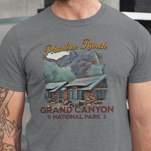 May include: Gray t-shirt featuring a graphic of Phantom Ranch in the Grand Canyon National Park. The design includes a rustic cabin illustration with the text "Phantom Ranch" in a decorative font above and "Grand Canyon National Park" below.