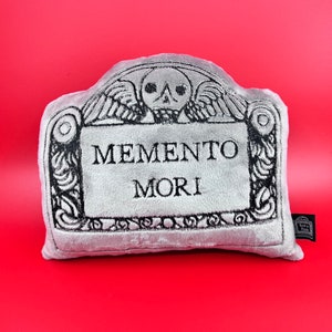 May include: A gray plush pillow shaped like a tombstone with the words "Memento Mori" embroidered on it.