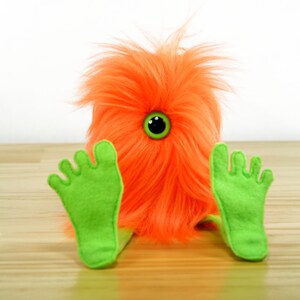 May include: A bright orange fuzzy monster with a single green eye sits with its green felt feet outstretched.