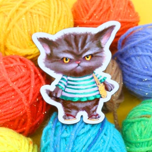 May include: A grumpy-looking gray cat wearing a green and white striped shirt with a yellow bag slung over its shoulder. The cat is surrounded by colorful yarn balls.