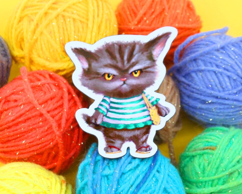 May include: A grumpy-looking gray cat wearing a green and white striped shirt with a yellow bag slung over its shoulder. The cat is surrounded by colorful yarn balls.