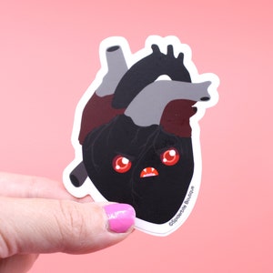 May include: A black heart sticker with red eyes and a menacing expression. The sticker is outlined in white and has the text "Spiderbite Boutique" in the bottom right corner.