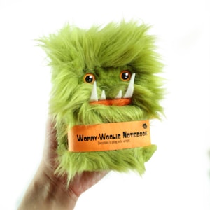 May include: Green fuzzy plush notebook cover with a toothy grin and orange eyes. The cover has a bright orange band with the text "Worry Woolie Notebook" and the tagline "Everything is going to be alright."
