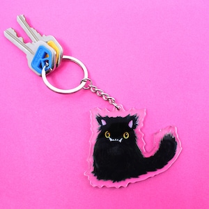 Puede incluir: Un llavero de gato negro con ojos amarillos y una cadena plateada. El llavero está unido a un juego de llaves con detalles azules y amarillos.