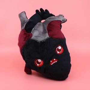 May include: A plush heart-shaped toy with a black, gray, and red fabric exterior. The toy has a cartoon-like face with red eyes and a toothy grin.