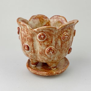 May include: A small, scalloped ceramic planter with a mottled brown and tan glaze. The exterior features raised circular accents with cross markings. The planter sits on three short feet and includes a matching circular saucer base.