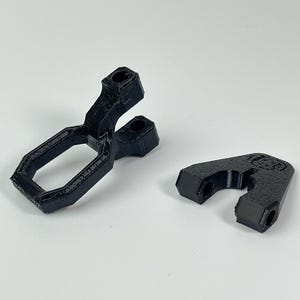 May include: Two black 3D-printed plastic components. One is a bracket with a rectangular aperture and two cylindrical projections. The other is a smaller, curved piece with a similar design, featuring a logo.