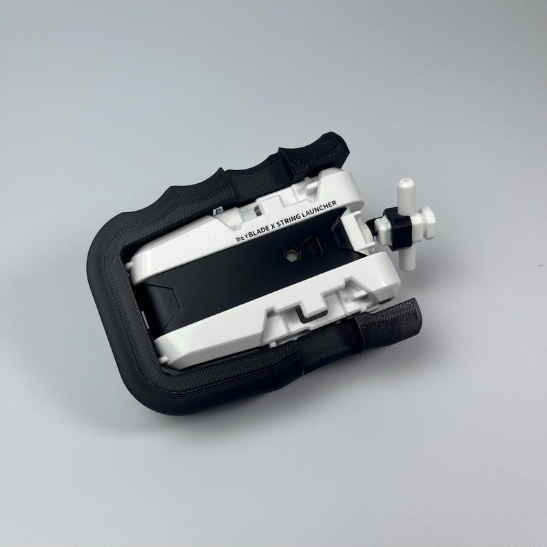 Puede incluir: Lanzador de cuerda Beyblade X blanco y negro con un mango negro. El lanzador tiene un mango blanco y una secci&oacute;n central negra. El texto "Beyblade X String Launcher" est&aacute; impreso en el lateral.