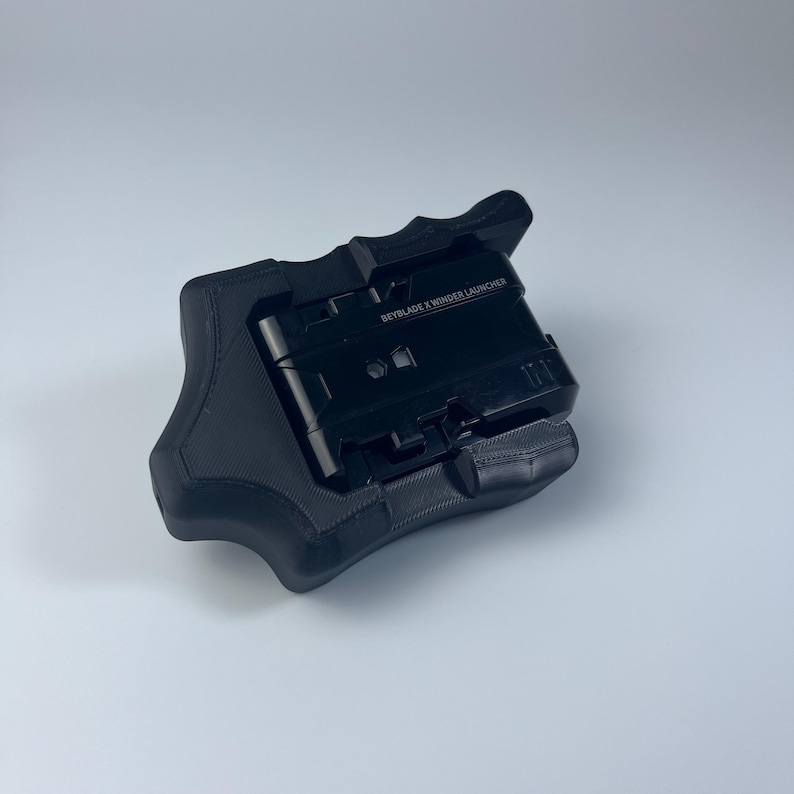 Puede incluir: Un lanzador negro Beyblade X Winder, un dispositivo port&aacute;til con un agarre texturizado. El lanzador tiene el texto "BEYBLADE X WINDER LAUNCHER" impreso en la parte superior. El dispositivo est&aacute; dise&ntilde;ado para lanzar peonzas Beyblade X.