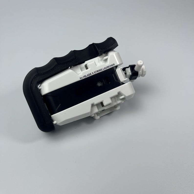 Puede incluir: Lanzador de cuerda Beyblade X blanco y negro con un asa negra. El texto "Beyblade X String Launcher" est&aacute; impreso en el lateral. El lanzador tiene una peque&ntilde;a boquilla blanca y negra en el extremo.