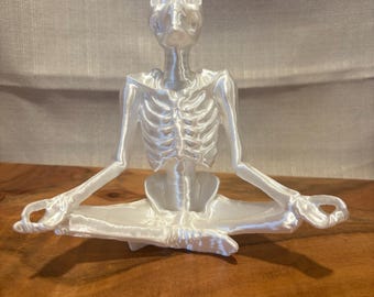 Può includere: Figurina di scheletro in plastica bianca seduta in una posa yoga con le gambe incrociate e le mani in mudra.