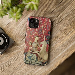 Puede incluir: Una funda de teléfono con un diseño de tapiz que presenta a una mujer, un unicornio y elementos florales sobre un fondo rojo. La funda está sobre una superficie de madera, con una cámara y vegetación en el fondo.