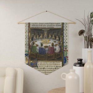 Op de afbeelding: Een middeleeuwse wandtapijt met een gedetailleerde illustratie van een ronde tafel met figuren in sierlijke kleding. Het kunstwerk heeft een crèmekleurige achtergrond met bloemenranden en tekst aan de boven- en onderkant. Het wandtapijt is opgehangen aan een houten staaf.
