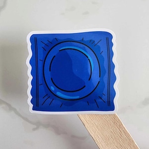 May include: A blue sticker featuring a stylized illustration of a condom wrapper. The design includes a central circular shape and radiating lines, all in dark blue against a lighter blue background. The sticker has a white scalloped border.
