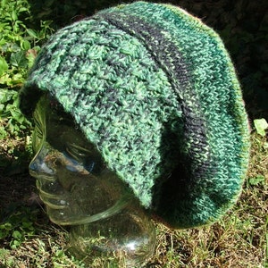 May include: A green and black knitted beret with a textured pattern. The beret is on a glass head form.