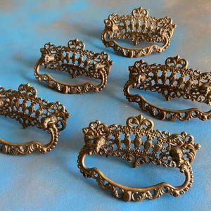 May include: Five antique bronze drawer pulls with ornate, filigree designs. Each pull features a curved handle and decorative embellishments, including floral and face motifs. The pulls are arranged on a blue surface.