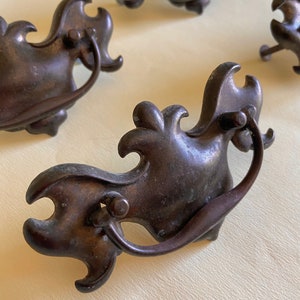 May include: Four antique bronze drawer pulls with ornate, decorative, scroll-like designs. Each pull has a loop handle.