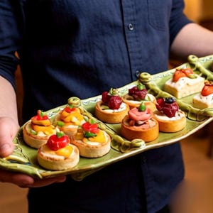 May include: A green, oblong serving tray holds an assortment of miniature tarts. The tarts are topped with colorful fruits and vegetables, including red peppers, strawberries, and blueberries. The tray is being held by a person wearing a dark blue shirt.