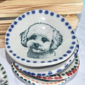 May include: A collection of small, hand-painted ceramic dishes featuring dog portraits. The dishes have various border designs and colors, including blue, green, and orange. Some dishes are stacked, and one contains wrapped candies.