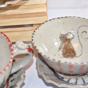 Puede incluir: Tazas de t&eacute; y platillos de cer&aacute;mica hechos a mano. Las tazas tienen un acabado moteado blanquecino con borde y asa rojos. Una taza presenta una ilustraci&oacute;n de rat&oacute;n marr&oacute;n. Los platillos tienen un borde festoneado con detalles rojos y azules.