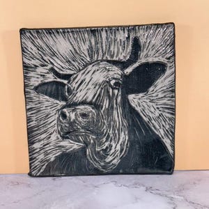 May include: A square ceramic tile featuring a black and white sgraffito illustration of a cow head. The design uses contrasting etched lines to create a textured effect, with the cow centered against a radiating pattern of light and dark strokes.