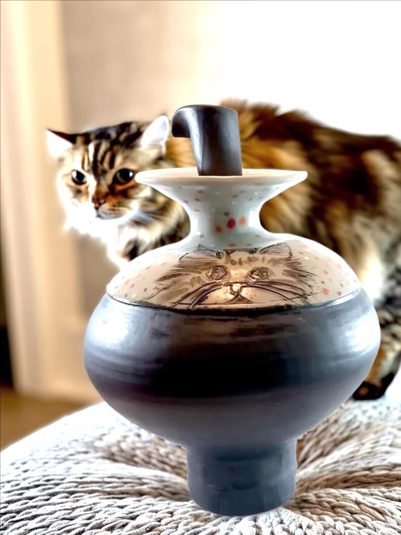 May include: A ceramic jar with a dark blue base and a cream-colored top featuring a cat face design. The lid has a dark handle. The jar is on a textured surface, with a blurred cat in the background.
