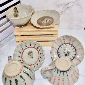 Puede incluir: Una colecci&oacute;n de vajilla de cer&aacute;mica hecha a mano, que incluye tazas y platillos. Las piezas presentan dise&ntilde;os &uacute;nicos pintados a mano con una paleta de colores neutros y acentos en rojo, verde y azul. El juego incluye un cuenco con una ilustraci&oacute;n de rat&oacute;n.