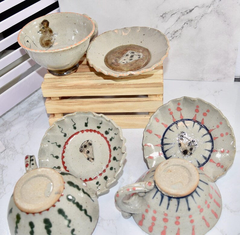 Puede incluir: Una colecci&oacute;n de vajilla de cer&aacute;mica hecha a mano, que incluye tazas y platillos. Las piezas presentan dise&ntilde;os &uacute;nicos pintados a mano con una paleta de colores neutros y acentos en rojo, verde y azul. El juego incluye un cuenco con una ilustraci&oacute;n de rat&oacute;n.