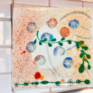 May include: A square fused glass tile featuring a white base with orange speckled accents. The design includes several circular blue and orange glass elements, a green vine-like pattern, and a thin green line along the bottom edge.