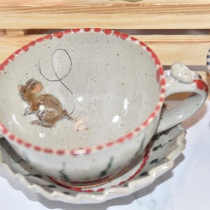 Puede incluir: Juego de taza y platillo de t&eacute; de cer&aacute;mica hechos a mano. La taza presenta una ilustraci&oacute;n caprichosa de un rat&oacute;n y un borde rojo. El platillo tiene un borde festoneado. El asa tiene un detalle en forma de rat&oacute;n. El juego es blanquecino con detalles marrones y rojos.