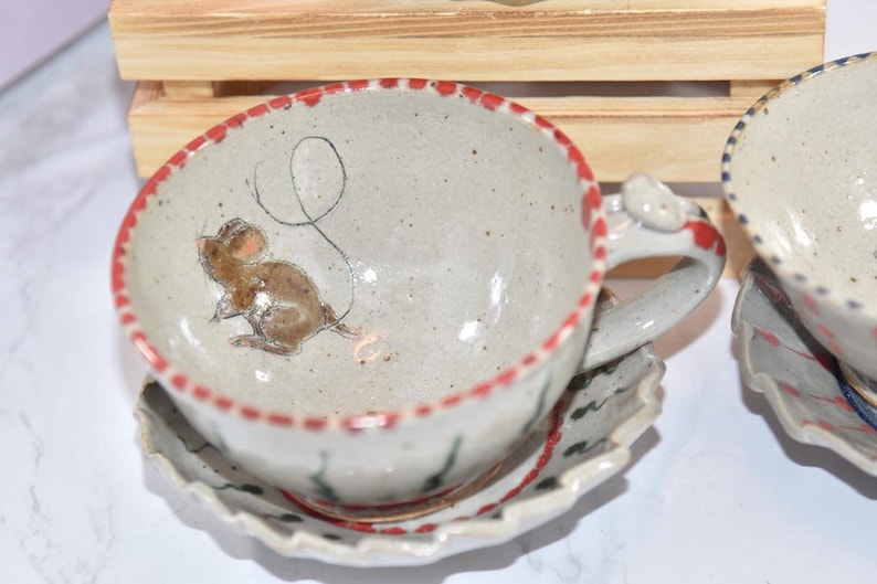 Puede incluir: Juego de taza y platillo de t&eacute; de cer&aacute;mica hechos a mano. La taza presenta una ilustraci&oacute;n caprichosa de un rat&oacute;n y un borde rojo. El platillo tiene un borde festoneado. El asa tiene un detalle en forma de rat&oacute;n. El juego es blanquecino con detalles marrones y rojos.