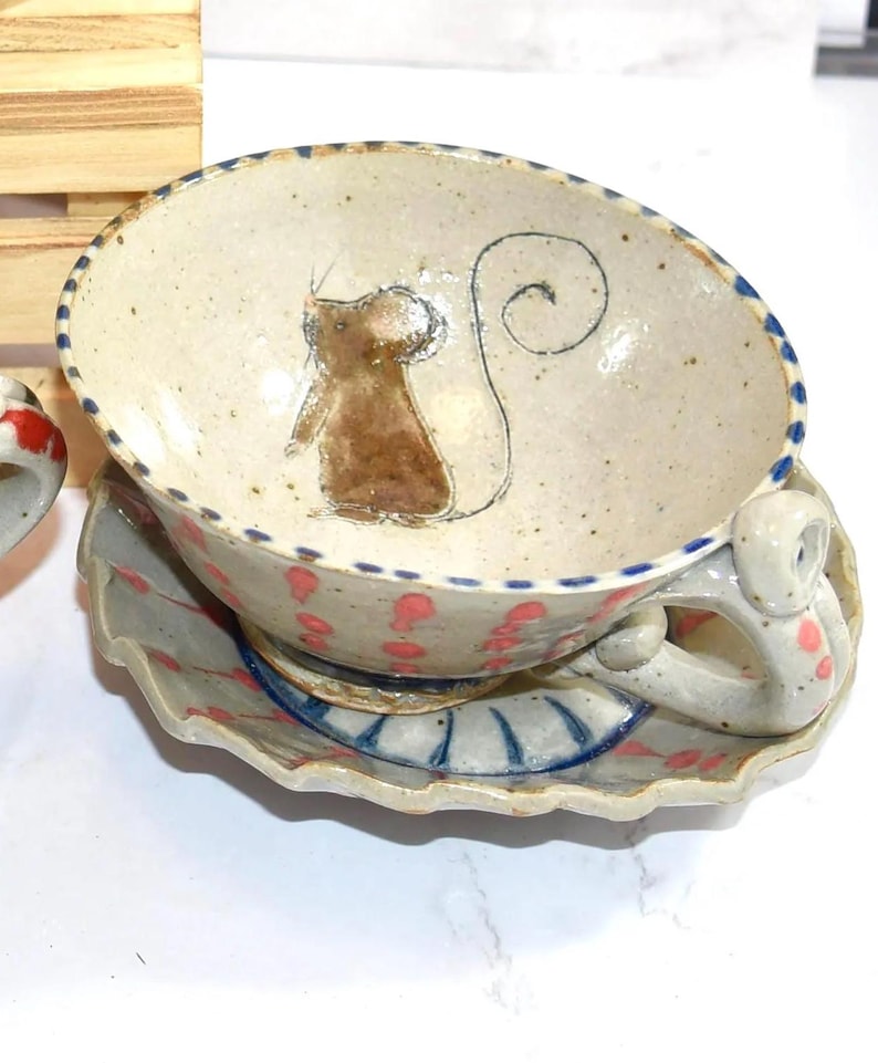Puede incluir: Juego de taza y platillo de t&eacute; de cer&aacute;mica pintados a mano. La taza presenta una ilustraci&oacute;n de rat&oacute;n marr&oacute;n y un borde punteado azul. El platillo tiene un borde festoneado y lunares rosas. El asa tiene una forma &uacute;nica.