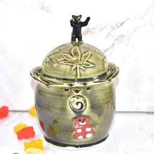 May include: A ceramic jar with a lid, featuring a black bear figurine on top. The jar is olive green with a maple leaf design on the lid and a red bear applique on the front. The jar is decorated with a spiral and small bear faces.