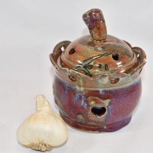 以下が含まれることがあります： 蓋付きの丸い釉薬陶器製ガーリックキーパー。蓋にはつまみがあり、容器全体は紫、茶、青のまだら模様で、縁はスカラップ状になっています。上部と側面には通気孔があり、横にはニンニクの球根が置かれています。