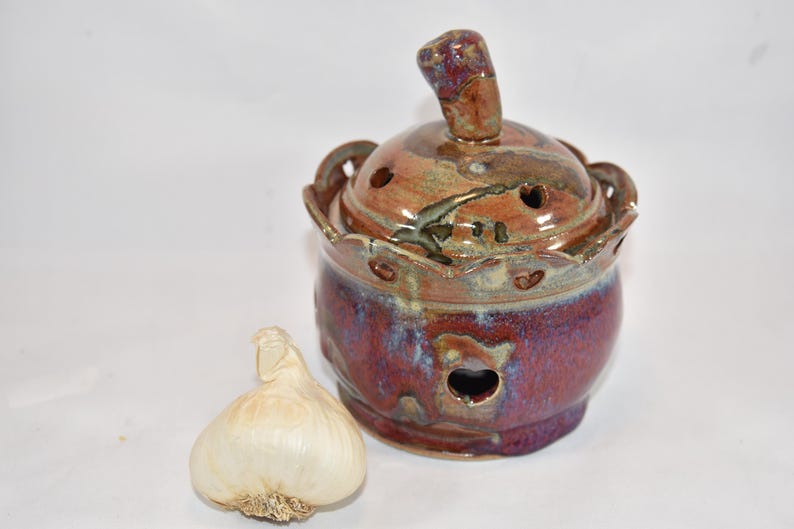 以下が含まれることがあります： 蓋付きの丸い釉薬陶器製ガーリックキーパー。蓋にはつまみがあり、容器全体は紫、茶、青のまだら模様で、縁はスカラップ状になっています。上部と側面には通気孔があり、横にはニンニクの球根が置かれています。