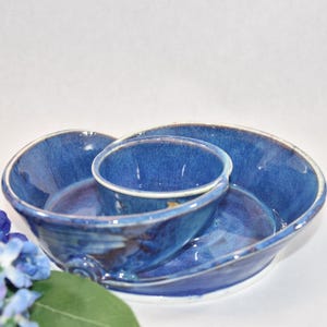 May include: A blue ceramic serving dish with a smaller bowl in the center. The dish has a unique shape and a speckled glaze.