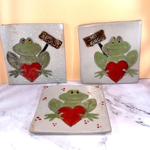 May include: Three ceramic tiles featuring green frogs holding red hearts. Two tiles have signs that read "RESIST" and "NO KINGS". The tiles are square with a speckled, off-white background. The frog illustrations are hand-painted.