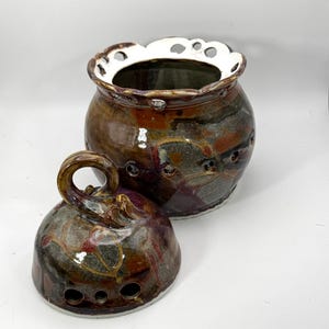 以下が含まれることがあります： 茶色、ベージュ、紫の斑点模様が施された、蓋付きの丸い陶器製の瓶。瓶の口は白い縁取りのスカラップ状になっており、本体と蓋には小さな円形の切り抜きが施され、蓋にはループ状の持ち手が付いています。