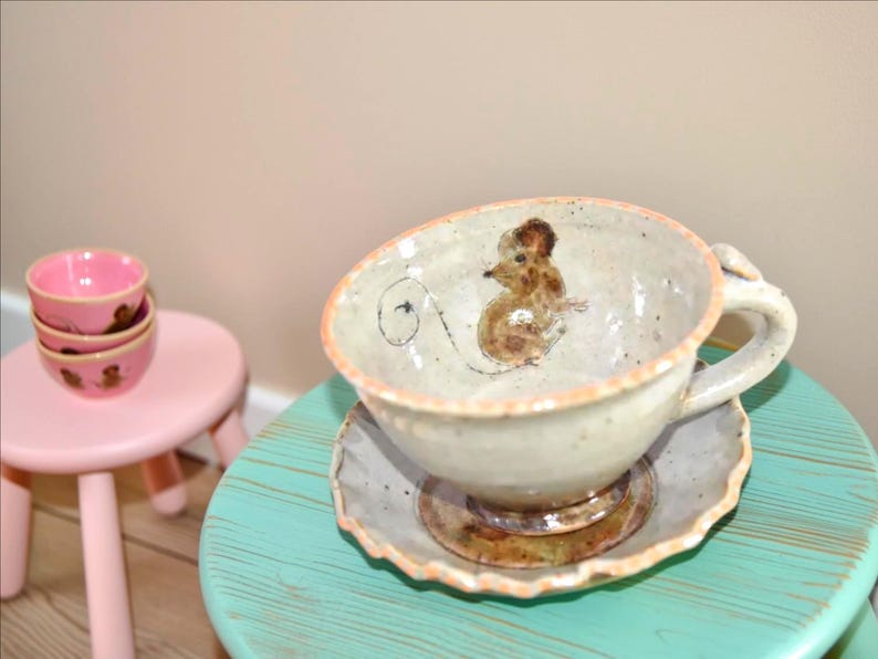 Puede incluir: Un juego de taza y platillo de t&eacute; de cer&aacute;mica hecho a mano con una ilustraci&oacute;n de rat&oacute;n. La taza es de color blanquecino con un dise&ntilde;o de rat&oacute;n marr&oacute;n y un borde naranja claro. El platillo tiene un dise&ntilde;o a juego. Tambi&eacute;n se ve una pila de tres cuencos rosas con ilustraciones de ratones.