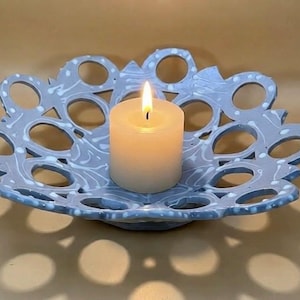 May include: A lit, white pillar candle sits in a decorative, light blue, openwork ceramic bowl. The bowl has a scalloped edge and a white, swirled pattern. The candle flame is bright against the soft background.