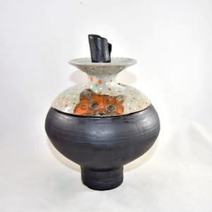 May include: A ceramic pet memorial urn with a black base and a light gray top decorated with orange polka dots. The lid features an orange cat face. A small black stopper fits into the top of this urn. The jar is a unique piece of art.